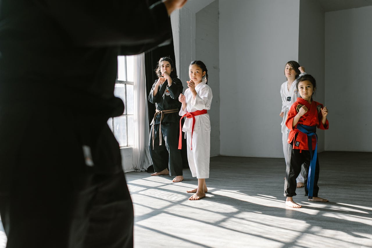 Group of children practicing martial arts indoors, guided by an instructor, in a sunlight-lit dojo.
